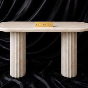 May include: An oval console table made of off-white marble with a smooth top surface and two cylindrical, vertically fluted legs. A small rectangular gold-colored plaque with the text AGATECRAFTSIND is centered on the table surface.