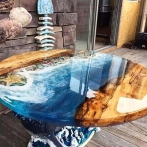 Op de afbeelding: Een ronde houten tafel met een blauw en wit epoxyhars blad dat lijkt op oceaan golven. De tafel is gemaakt van natuurlijk hout met een rustieke afwerking.