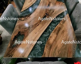 Puede incluir: Una gran mesa de comedor de madera con una incrustación de resina epoxi verde y azul. La mesa está rodeada de sillas grises y un cojín gris. La mesa está en una habitación con plantas verdes.