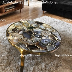 May include: A round, gold-toned coffee table with a top made of agate slices in shades of brown, grey, and white. The table has three legs and is sitting on a fluffy white rug.