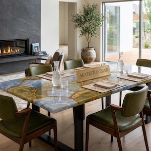 Op de afbeelding: Een rechthoekige eettafel met een gepolijst oppervlak van blauwe en gouden agaatstenen plakken. De tafel heeft een zwart metalen onderstel en is omringd door zes olijfgroene stoelen. Een houten bordje met de tekst AGATECRAFTSIND staat op de tafel.