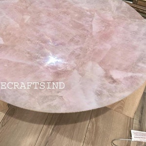 May include: A round, light pink rose quartz table with a light wood base. The table's surface is made of multiple pieces of rose quartz. The text "AGATECRAFTSIND" is visible on the table. The table is approximately 24 inches in diameter.