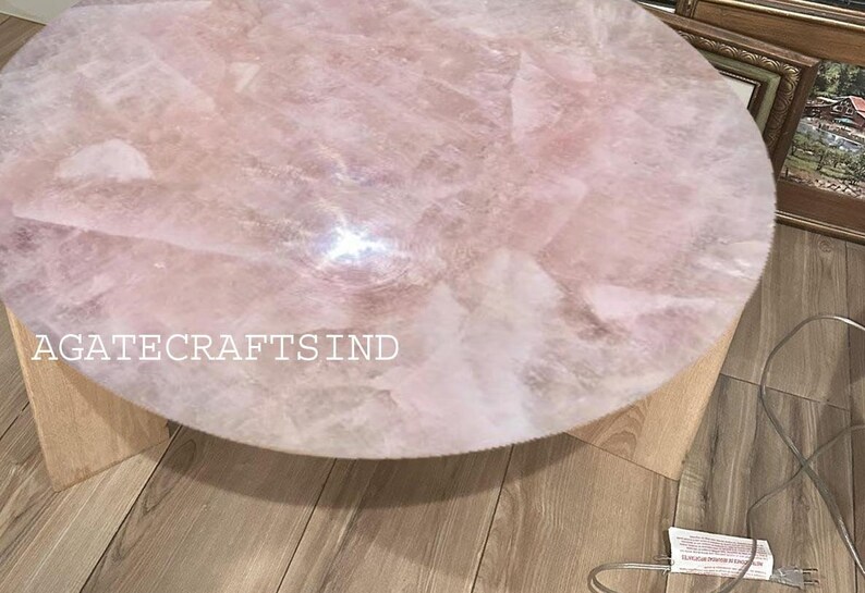 May include: A round, light pink rose quartz table with a light wood base. The table's surface is made of multiple pieces of rose quartz. The text "AGATECRAFTSIND" is visible on the table. The table is approximately 24 inches in diameter.