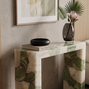 Op de afbeelding: Een consoletafel opgebouwd uit rechthoekige stukken groen en wit steen met een gepolijste afwerking. Bovenop ligt een beige boek met de titel Mid-Century Marvels, een matzwarte kom en een donkere glazen vaas met een roze bloem en groene bladeren.