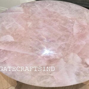 May include: Round coffee table with a light pink rose quartz top. The surface is made of irregular pieces of the stone, creating a unique pattern. The table has a light wood base and the text "AGATECRAFTSIND" is visible.