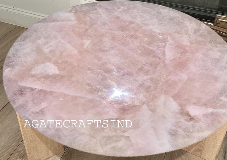 May include: Round coffee table with a light pink rose quartz top. The surface is made of irregular pieces of the stone, creating a unique pattern. The table has a light wood base and the text "AGATECRAFTSIND" is visible.