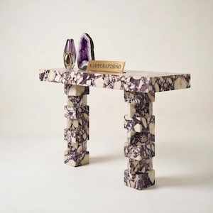 May include: A rectangular table with a purple and white patterned top and stacked block legs. The table holds a purple vase, a geode, and a gold sign that reads "AGATE CRAFTS IND".