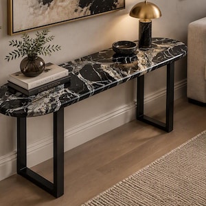 Op de afbeelding: Een consoletafel met een langwerpig, ovaal blad van zwart marmer met opvallende witte aders. De tafel wordt ondersteund door twee rechthoekige, matzwarte metalen poten. Op het oppervlak staan een kleine glazen vaas, twee boeken en een schaal.