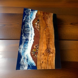 Könnte beinhalten: Ein einzigartiges Holzkunstwerk mit einer Strandszene. Das Kunstwerk kombiniert einen Abschnitt aus Naturholz mit einem Harzdesign, das Meereswellen, Sand und Kieselsteine darstellt. Die Wellen sind weiß und blau, der Sand ist rotbraun und die Kieselsteine sind mehrfarbig.