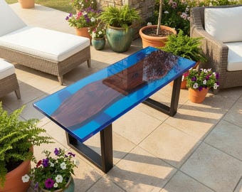 May include: A rectangular outdoor coffee table with a blue resin top and a dark wood inlay. The table has black metal legs and is surrounded by potted plants and patio furniture. The table measures approximately 48 inches long.