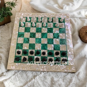 Pode incluir: Um tabuleiro de xadrez verde e branco feito de madrepérola e malaquita incrustados. O tabuleiro está configurado para uma partida de xadrez com peças pretas e brancas.