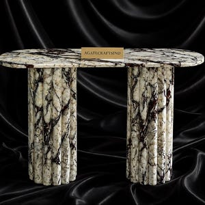 May include: An oval console table crafted from white marble with dark brown veining. The table features a smooth top and two cylindrical, fluted pedestal bases. A small rectangular sign with the text AGATECRAFTSIND sits on the tabletop.
