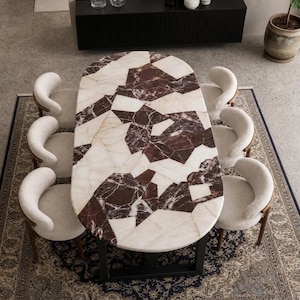 Può includere: Un tavolo da pranzo ovale con un piano a mosaico composto da pezzi di marmo bianco e marrone scuro, sostenuto da una struttura in metallo nero. Sei sedie curve con rivestimento testurizzato color crema e gambe in legno circondano il tavolo.