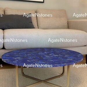 May include: A round coffee table with a blue lapis lazuli top and a gold metal frame. The table is in front of a white couch with gray pillows.