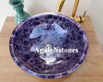 May include: A round, purple and white agate stone sink with the text "AgateNstones" in white. The sink is on a wooden surface, with a gold faucet and a teal vase in the background.