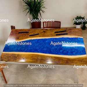 May include: A large wooden table with a blue epoxy river running through the center. The table has a natural wood finish and is supported by a metal base.