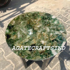 May include: A round green agate table top with a black metal base. The table top is made of polished agate with a variety of green shades and patterns. The text "AGATECRAFTSIND" is visible on the table top.