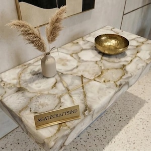 May include: A rectangular console table featuring a white agate stone surface with gold-toned veining. A small beige vase with dried pampas grass and a hammered gold bowl sit on top. A gold-colored sign displays the text AGATECRAFTSIND.