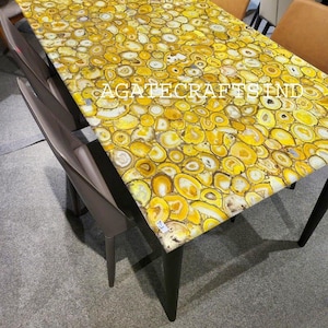 May include: A rectangular table with a yellow agate stone top. The surface is composed of numerous circular and oval-shaped yellow and white agate slices. The table has dark legs and is surrounded by chairs. The text "AGATE CRAFTS IND" is visible on the table.