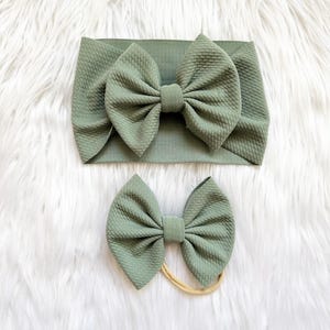 May include: Two-piece set of sage green baby headbands. One is a wide, textured headband with a large bow. The other is a smaller bow on a thin, neutral-colored elastic band. The headbands are displayed on a fluffy white surface.