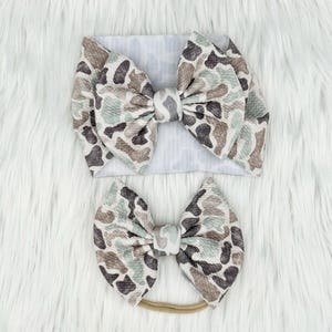 May include: Two-piece set of baby headbands. One is a wide headband with a large bow, and the other is a smaller bow on a thin elastic band. Both bows feature a camouflage pattern in shades of brown, gray, and green.