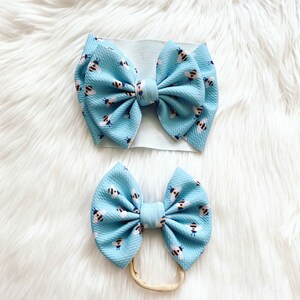 May include: Two blue hair bows with a bee print. The larger bow is on a white headband and the smaller bow is on a nude nylon headband.