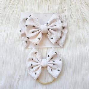 May include: Two white baby headbands with a repeating black bee pattern. One headband is a wide band, and the other is a smaller bow on a thin, beige elastic. The headbands are on a white, fluffy surface.