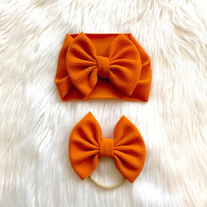 May include: Two orange fabric headbands with large bows. The larger headband is a knotted headband and the smaller headband is a hair tie.