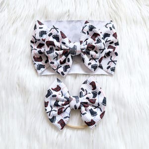 May include: A white headband and bow set featuring a repeating pattern of hockey skates and crossed hockey sticks in black, red, and gray. The bows are made of a textured fabric.