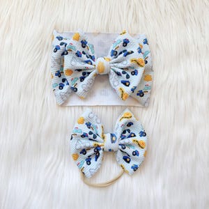 May include: Two baby bows, one on a headband and one on a nylon band. Both bows are light blue with a pattern of blue, yellow, and white vehicles. The headband bow is larger than the nylon band bow.