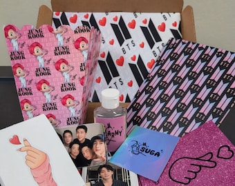 May include: A brown cardboard box filled with BTS-themed merchandise. Items include pink and white patterned paper, photo cards, a hand sanitizer bottle, and stickers. The box is decorated with hearts and the BTS logo.