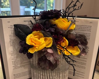 Puede incluir: Arreglo floral decorativo con flores amarillas y moradas, detalles en negro y un elemento decorativo plateado. Las flores están dispuestas en un jarrón hecho con páginas de libro dobladas.