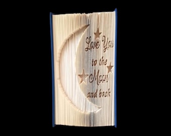 Può includere: Un libro con una copertina blu è stato piegato in un design tridimensionale di una luna crescente e stelle. Il libro ha il testo "Love you to the Moon and back" inciso sulle pagine.