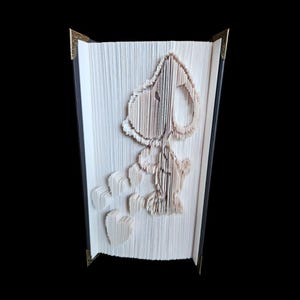 May include: A handmade book art sculpture featuring Snoopy from the Peanuts comic strip. The artwork is created by meticulously folding the pages of a book to form the character's image. The book has a black spine and gold corner protectors.
