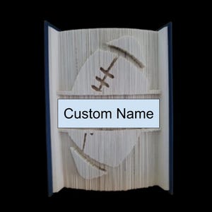 May include: A white book with a football design cut out of the pages. The book is open and the football design is visible. The text "Custom Name" is printed on a blue rectangle.