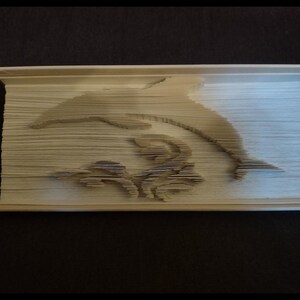 May include: A white book with pages folded into a 3D sculpture of a dolphin jumping out of the water. The dolphin is in the center of the book and is facing the right side of the image. The dolphin is surrounded by a design of stylized waves.
