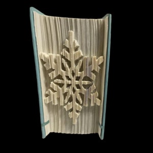 Op de afbeelding: Een boek met de pagina's gevouwen in de vorm van een sneeuwvlok. De sneeuwvlok is wit en het boek is bedekt met een lichtblauwe stof.