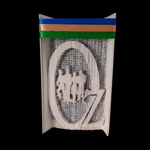 Puede incluir: Una escultura de arte de libro de la letra "O" con una silueta de los personajes de El mago de Oz en el interior. El libro está encuadernado en papel marrón y tiene una cinta verde, dorada y azul atada alrededor.