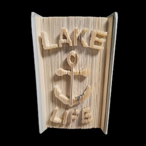 Pode incluir: Escultura de livro com as palavras "LAKE LIFE" e uma âncora cortada nas páginas. O livro está aberto, com o texto e o desenho da âncora visíveis nas páginas. O livro está sobre um fundo preto.