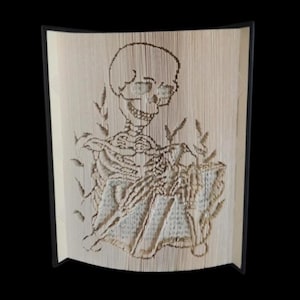 May include: A book art sculpture of a skeleton sitting and reading a book. The sculpture is made from the pages of a book that have been carefully cut and folded to create the image.