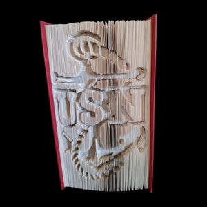 Puede incluir: Un libro con las palabras "USN" dobladas en las p&aacute;ginas, con un dise&ntilde;o de ancla debajo. El libro es rojo por fuera.