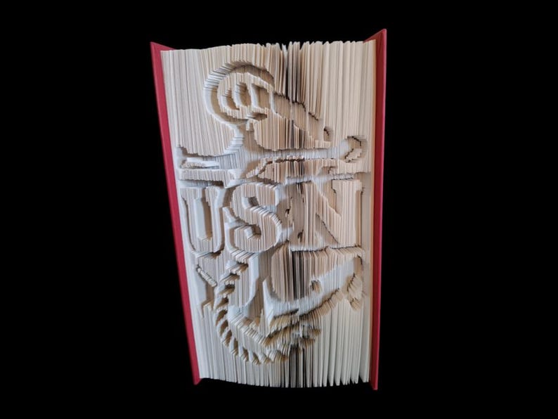 Puede incluir: Un libro con las palabras "USN" dobladas en las p&aacute;ginas, con un dise&ntilde;o de ancla debajo. El libro es rojo por fuera.