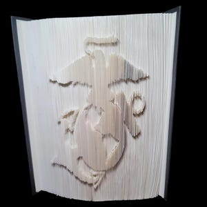 Puede incluir: Un libro blanco con el emblema del Cuerpo de Marines doblado en las p&aacute;ginas. El emblema es un &aacute;guila estilizada, un globo terr&aacute;queo y un ancla.