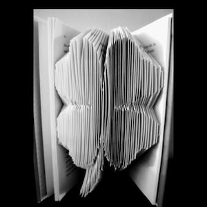 May include: Black and white photograph of a book with pages folded to create a four-leaf clover shape. The clover is centered, with the book open to display the intricate paper folding. The background is black.