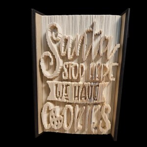 Op de afbeelding: Een boeksculptuur met de woorden "Santa Stop Here We Have Cookies" uitgesneden in de pagina's. De tekst is in een grillige lettertype en het boek is open, waardoor de ingewikkelde sneden zichtbaar zijn. De achtergrond is zwart.