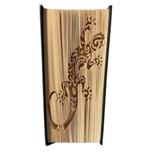 May include: A decorative book with a lizard design carved into the pages. The lizard is brown and has intricate details. The book has a black spine and the pages are a light tan colour.