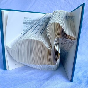 May include: A book with pages folded into a 3D shape resembling a bird. The book is open and the folded pages are visible. The book has a teal blue cover.