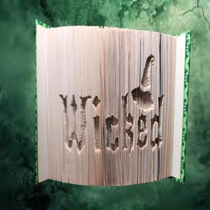 May include: A book with the word "Wicked" cut out of the pages. The book is open and the pages are folded to create a 3D effect. The book is on a green background.