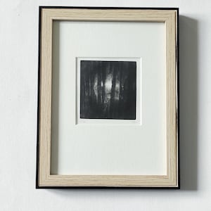 Peut inclure: Une estampe encadrée en noir et blanc représentant une scène de forêt. L'œuvre d'art carrée est encadrée d'un cadre en bois clair et d'un bord intérieur noir. L'estampe montre des lignes verticales suggérant des arbres, avec une zone lumineuse au centre.