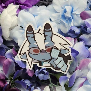 May include: A die-cut sticker featuring a cartoon character with blue fur, red eyes, and brown striped ears. The sticker is surrounded by a variety of artificial flowers in shades of purple, white, and blue.